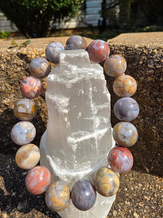Alashan Agate bracelet featuring large round beads with swirling pastel shades of pink, purple, beige, grey and gold draped around a selenite tower on a natural stone background. This nurturing stone encourages balance, creativity and stability.