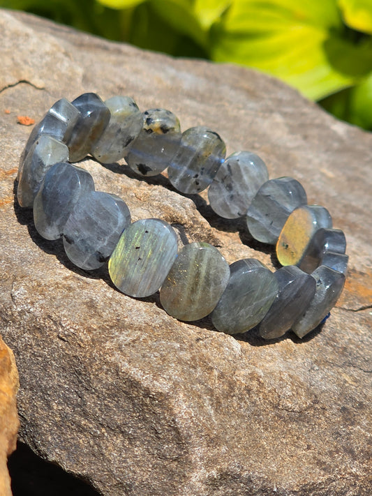 Faceted labradorite bracelet featuring thick disc-shaped beads with a gray base and flashes of blue and green labradorescence, 9–10 mm, displayed on a rock with greenery; a stone of transformation and intuition.