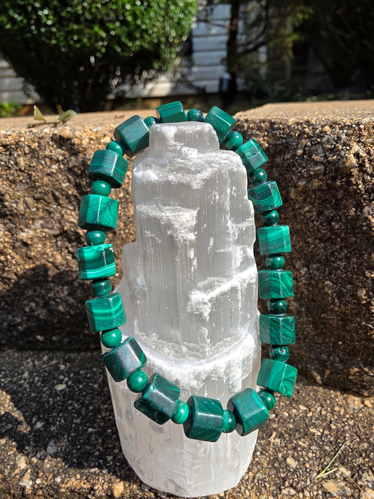 Malachite cylinder bracelet featuring cylindrical malachite beads in deep green hues with natural banding, draped around a selenite tower on a stone surface and greenery; malachite fosters transformation, protection, and healing energy.