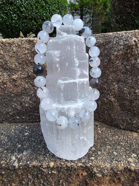 Rutilated quartz bracelet featuring clear beads with black rutile needles, displayed on a selenite tower; this gemstone is believed to amplify clarity and energy.