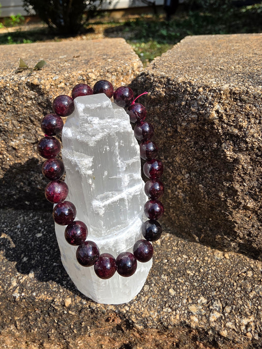 Starlight Garnet bracelet featuring round 10mm deep red garnet beads wrapped around a selenite tower on a stone surface; this stone inspires passion, courage, and grounding energy.