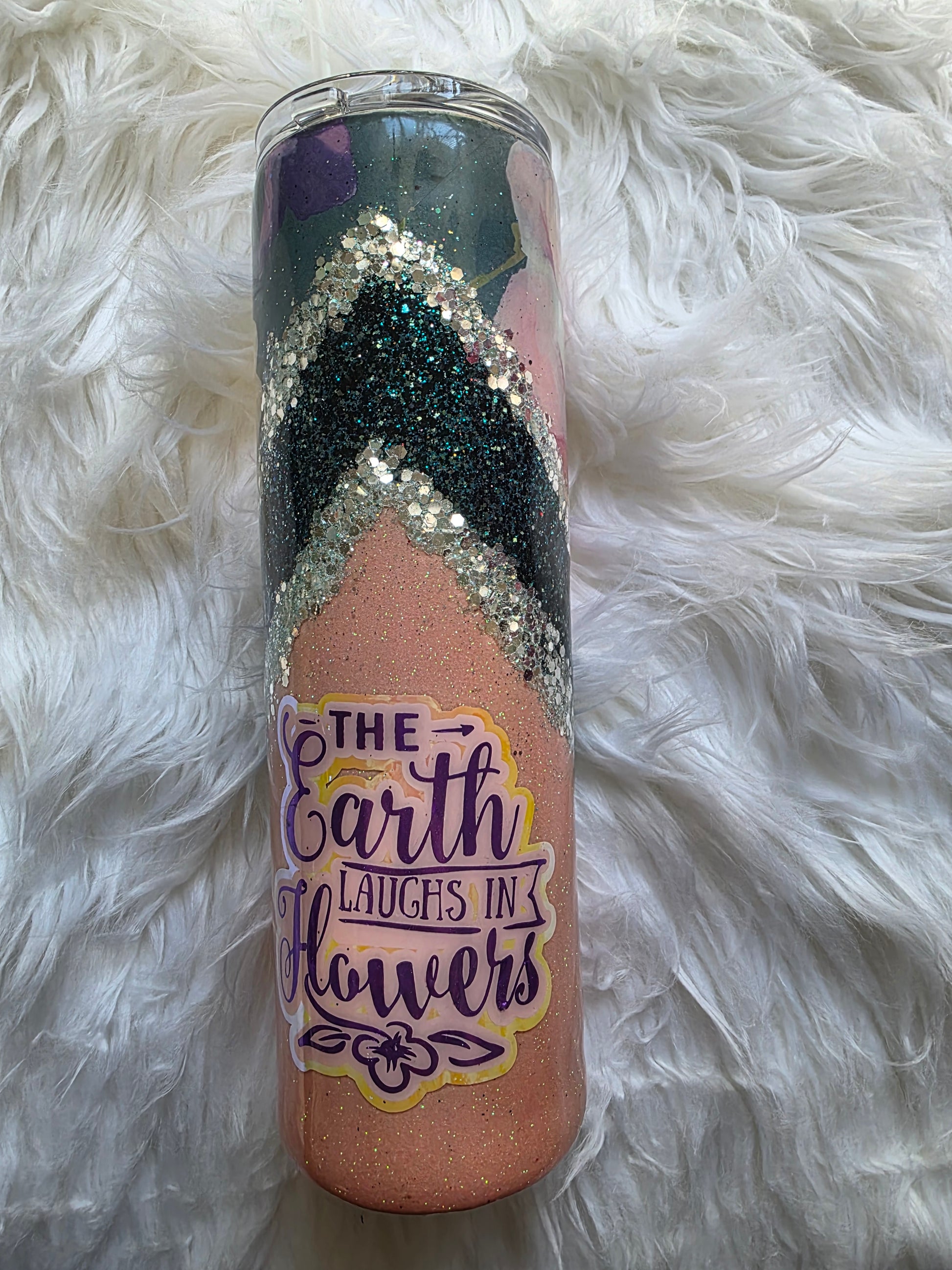Epoxy tumbler showing the quote ‘The Earth Laughs in Flowers’ in purple script on a glittery peach and teal background.